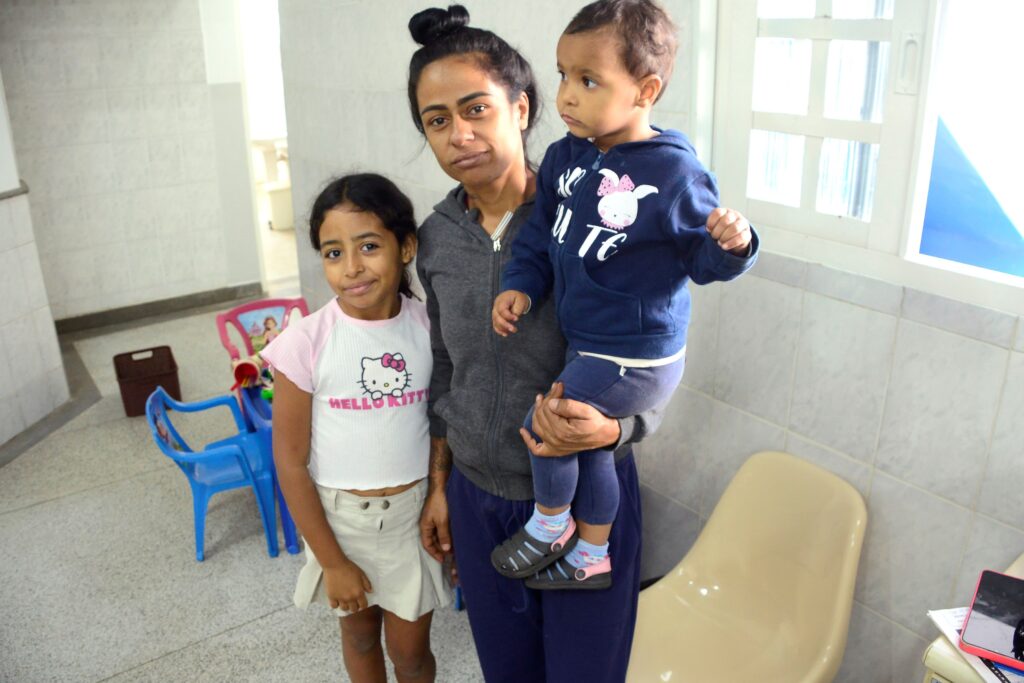 Foto interna mostrando três pessoas próximas umas das outras em um ambiente com paredes e piso em azulejo claro — parece uma sala de espera ou área de acolhimento. Pessoas: mulher jovem ao centro segurando uma criança pequena no braço direito e com uma menina mais velha apoiada à sua esquerda. Expressões e postura: a mulher tem expressão séria/neutra, cabelo preso em coque; a menina mais velha sorri levemente; a criança pequena olha para a direita com expressão neutra. Roupas: a menina mais velha usa camiseta branca com estampa da Hello Kitty e saia clara; a mulher veste moletom cinza escuro e calça escura; a criança usa conjunto azul-marinho com capuz e sandálias escuras mais meias estampadas. Ambiente: cadeiras plásticas infantis azuis e um brinquedo no chão atrás deles; janela grande à direita deixando entrar luz natural; piso e paredes revestidos com azulejos claros; móveis simples (cadeira bege e mesa com celular e papéis no canto). Iluminação: luz natural forte vinda da janela, criando sombras suaves. Atmosfera: cena cotidiana, íntima e tranquila, possivelmente em local de atendimento ou visitação com espaço para crianças.