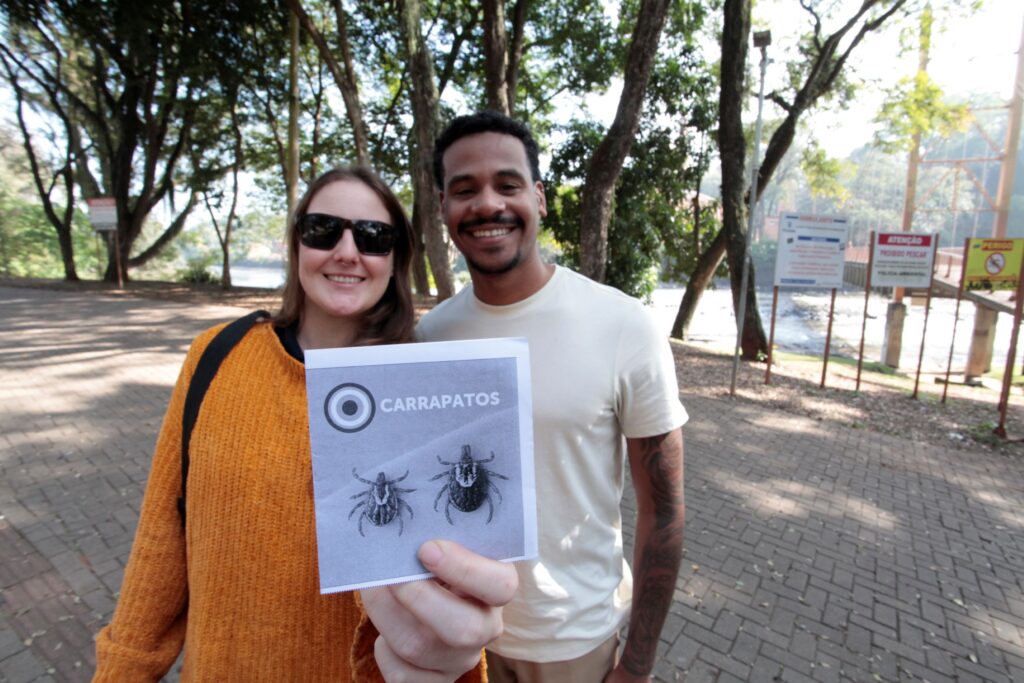 Dois adultos sorrindo em primeiro plano seguram (ou recebem) um folheto sobre “CARRAPATOS”; o folheto está bem próximo à câmera e parte dele aparece em foco. Pessoa à esquerda: aparenta ser mulher, usa suéter/poncho laranja, óculos escuros e uma mochila preta; sorri para a câmera. Pessoa à direita: aparenta ser homem, camiseta clara, barba curta e sorriso largo; tem tatuagens visíveis no braço direito. Mãos/folheto: uma mão à frente segura o folheto com desenhos de carrapatos e o título “CARRAPATOS”; os dedos estão em destaque no canto inferior da imagem. Ambiente: parque arborizado junto a um rio; piso de blocos e várias árvores altas ao fundo. À direita, placas de aviso e uma grade próximas à margem do rio. Iluminação e clima: dia ensolarado com sombras de árvores no chão; atmosfera descontraída e informativa, como de ação de conscientização ao ar livre.