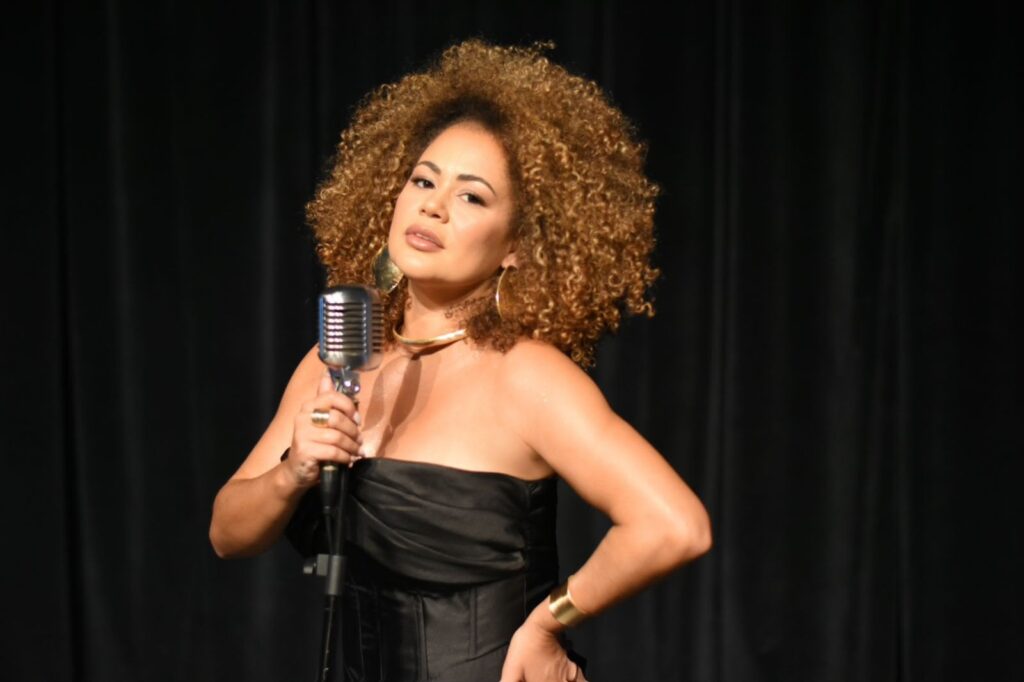 Mulher em primeiro plano, provavelmente artista/cantora em palco. Detalhes observáveis: Composição: enquadramento médio (do peito para cima) com fundo de cortina preta, foco nítido na pessoa. Aparência: pele morena clara, cabelo volumoso e cacheado em tom castanho-dourado; maquiagem discreta realçando olhos e lábios. Roupa e acessórios: vestido ou corpete preto sem alças; colar rígido dourado, brincos grandes, pulseira larga dourada e anel visível na mão que segura o microfone. Microfone e postura: microfone retro/analógico prateado apoiado em pedestal — ela segura o cabo/haste com a mão esquerda; postura confiante, braço direito na cintura e expressão serena/confiante, olhando levemente para a câmera. Iluminação e atmosfera: luz quente direcionada que destaca a pele e o brilho nos acessórios; cenário minimalista que sugere apresentação musical íntima (jazz/blues/soul).