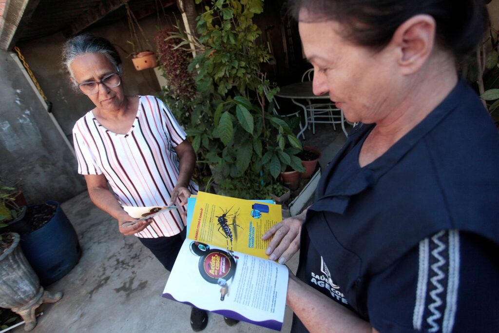 Uma agente de saúde comunitária conversa com uma moradora em uma área externa, entregando material informativo sobre a prevenção da proliferação do mosquito Aedes aegypti, em um momento de conscientização pública. A composição da cena é construída a partir de um ângulo em primeiro plano, onde a agente de saúde está posicionada à direita, dominando a visão com o folheto informativo aberto, enquanto a moradora está à esquerda, voltada para a agente. O enquadramento sugere uma interação direta e educacional, onde o foco principal recai sobre o livreto que contém ilustrações do mosquito. O posicionamento das figuras cria uma diagonal que conduz o olhar do espectador diretamente para o material educativo que está sendo compartilhado. A agente de saúde é vista de perfil, vestindo um colete azul escuro com detalhes brancos, onde se lê "AGENTE DE SAÚDE", denotando sua função profissional. Ela possui cabelos escuros presos. A moradora, posicionada em um ângulo ligeiramente superior, possui cabelos grisalhos curtos, usa óculos de grau e veste uma blusa listrada nas cores branca, preta e salmão. Sua expressão é de atenção e foco enquanto examina o documento. Ambas as figuras mantêm uma postura séria e engajada, refletindo a importância da orientação que está sendo transmitida durante a visita domiciliar. Esta é uma fotografia documental que prioriza o registro de uma ação cotidiana de saúde pública, sem grandes artifícios estilísticos. A execução é direta, com uma iluminação natural que provém de uma fonte externa, provavelmente luz do dia filtrada por uma varanda ou área coberta. As cores são naturais, com predominância dos tons de azul do uniforme e do amarelo vibrante do folheto, que se destaca visualmente como o elemento central de informação na cena, contrastando com o ambiente mais sóbrio e concreto do local de residência. O cenário é uma varanda ou área de serviço externa de uma casa, caracterizada pelo piso de cimento queimado e pela presença de vasos de plantas, incluindo uma planta ornamental em um vaso azul no canto esquerdo. A iluminação é difusa, vinda de cima e das laterais, típica de áreas de sombra próximas à entrada de residências, o que confere um tom sóbrio e realista à imagem. O ambiente reflete uma simplicidade comum em áreas residenciais, onde o encontro ocorre de maneira informal, mas focada na responsabilidade coletiva de combate a doenças, criando um clima de seriedade e diligência.