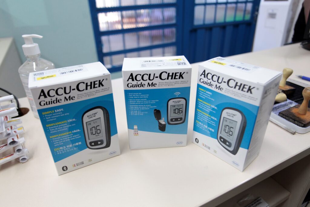 Três caixas do medidor de glicose Accu-Chek Guide Me alinhadas sobre um balcão claro, em primeiro plano. Cada embalagem mostra a imagem do aparelho com leitura “106 mg/dL”, fundo branco e faixa azul com instruções em vários idiomas. À esquerda há um frasco com pump de álcool em gel parcialmente visível; à direita aparecem carimbos e uma caneta. Ao fundo vê‑se uma grade azul/portão desfocada e uma parede clara. A iluminação é interna, uniforme, e a cena transmite um ambiente de farmácia ou clínica com produtos prontos para venda ou distribuição.