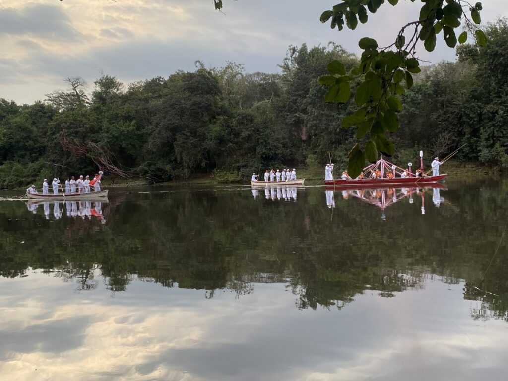 Foto de uma procissão ou cortejo sobre um rio ao entardecer. Principais elementos: Três embarcações alinhadas no centro-direita da imagem — duas maiores (especialmente a da direita, vermelha e decorada) e uma menor entre elas. Muitas pessoas vestidas predominantemente de branco nas canoas; algumas usam coletes laranja e outras seguram varas longas para manobrar as embarcações. Estandarte/estrutura decorativa sobre a canoa da direita, com fitas claras que se refletem na água. Reflexos nítidos das embarcações, pessoas e da vegetação no espelho d’água calmo, criando simetria horizontal. Margem arborizada densa ao fundo, em tons de verde-escuro, que contrasta com o céu claro. Céu parcialmente nublado com luz suave de fim de tarde, dando tom dourado/acinzentado à cena. Ramos e folhas em primeiro plano no canto superior direito, enquadrando a foto e acrescentando profundidade. Atmosfera geral: cerimoniosa e tranquila, com sensação de ritual ou celebração comunitária.