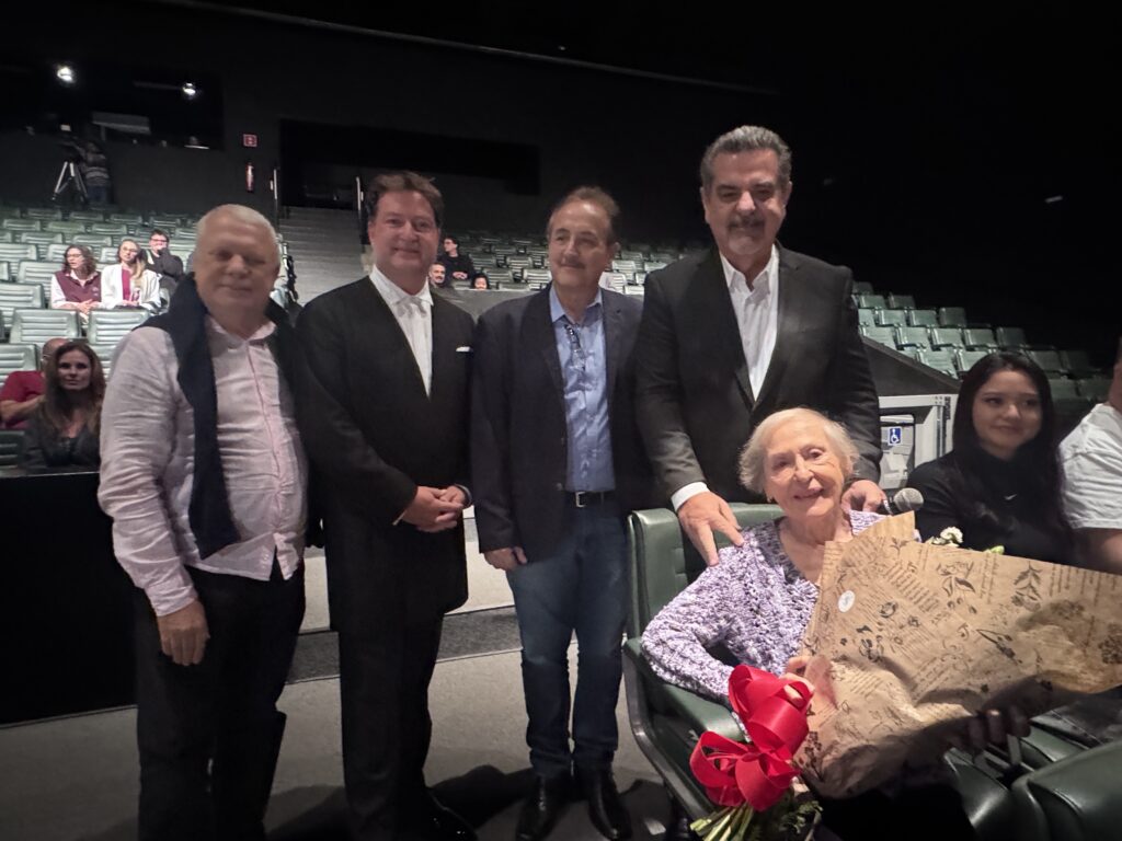 Nesta foto, um grupo de cinco pessoas posa para uma foto em um auditório ou sala de cinema. Em pé: À esquerda, um homem com camisa rosa clara e um suéter escuro jogado sobre os ombros. Ao lado dele, um homem de terno preto e camisa branca. No centro, um homem de blazer escuro, camisa azul e jeans. À direita, um homem alto de terno preto e camisa branca, com a mão no ombro da mulher sentada. Sentada: Uma senhora idosa, sorridente, segura um grande buquê de flores com um laço vermelho vistoso. Background: À direita, há uma jovem sentada observando, e ao fundo, vê-se a plateia do auditório com assentos verdes. A iluminação sugere um ambiente fechado, como um teatro ou cinema.
