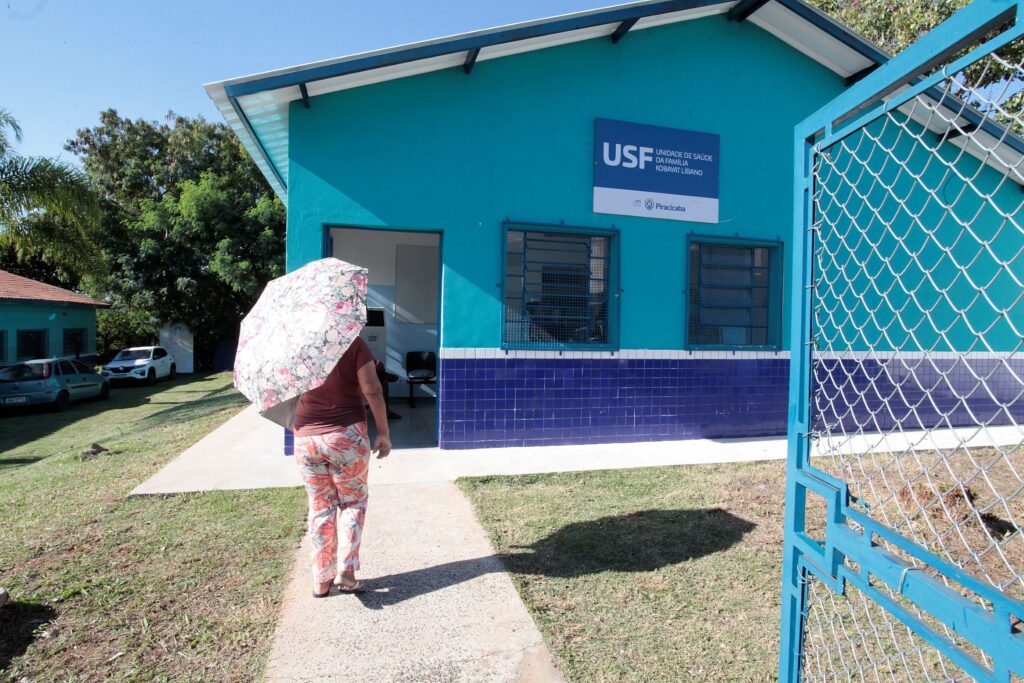 Prédio pequeno de unidade de saúde pintado de azul-turquesa com faixa de azulejos azuis escuros na base. Placa acima da janela com as letras grandes “USF” (Unidade de Saúde da Família) e logo/menção a Piracicaba. Estrutura: uma porta de entrada aberta e duas janelas com grades externas; telhado com beiral branco. Pessoa: uma pessoa de costas, segurando um guarda-sol florido rosa e branco, caminhando em direção à entrada — roupa com estampa floral e sandálias. Ambiente: gramado ao redor, calçada de concreto que leva à porta; ao fundo há árvores e alguns carros estacionados. Elementos: portão de grade azul à direita em primeiro plano; iluminação indica dia claro e ensolarado.
