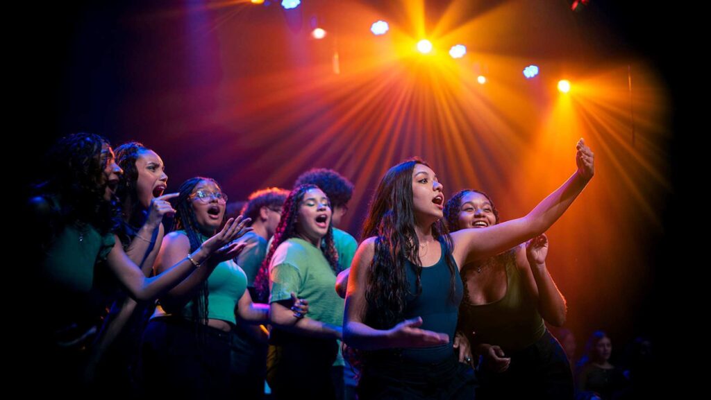 A imagem mostra um grupo de jovens se apresentando no palco, provavelmente um coral ou grupo de teatro musical.Detalhes da cena:Apresentação: São cerca de 7 jovens, maioria mulheres, vestindo roupas em tons de verde e azul. Elas estão cantando com muita expressão: boca aberta, mãos gesticulando, corpo envolvido. Destaque: A moça à direita, de regata azul-escura e cabelo longo, está com um braço levantado para o alto e parece liderar a música. As outras acompanham com gestos sincronizados, sorrisos e energia.Iluminação: O palco está iluminado por refletores fortes. A luz predominante é amarela/dourada, formando raios que descem do alto. Tem também luzes azuis e roxas nas laterais, criando contraste e clima de show. O fundo é escuro, o que destaca o grupo.Atmosfera: Dá pra sentir a emoção e a força da apresentação. Parece um momento de clímax da música, com todo mundo conectado e entregando a performance.O cenário passa alegria, juventude e talento. Parece uma apresentação de escola, projeto social ou grupo artístico.