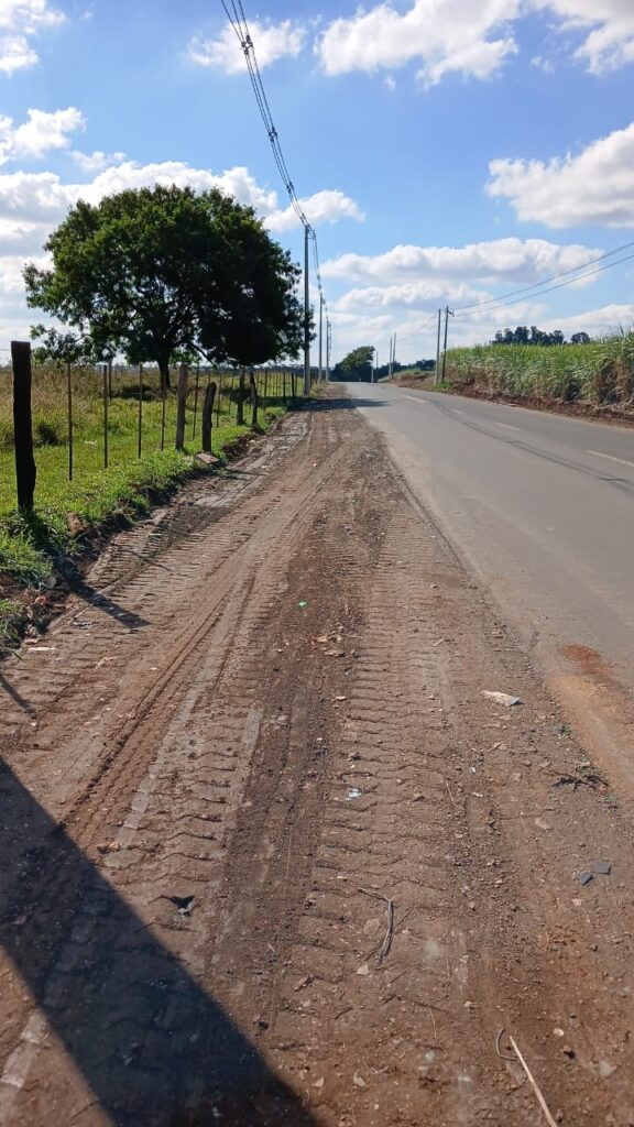 Esta imagem mostra o acostamento de uma estrada pavimentada em uma área rural. O solo no acostamento é de terra batida, marcado com rastros profundos de pneus ou esteiras de veículos pesados. À esquerda, há uma cerca de arame com postes de madeira e uma árvore grande. Do lado direito, ao lado da estrada asfaltada, existe uma plantação que parece ser de cana-de-açúcar. O dia está ensolarado, com um céu azul e poucas nuvens, e há uma linha de postes de energia elétrica seguindo o traçado da estrada ao longe.