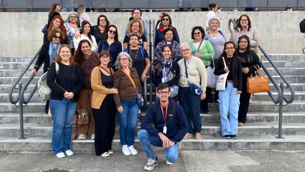 A imagem mostra um grupo de aproximadamente 25 pessoas reunidas para uma fotografia em uma escadaria de concreto, em frente a um prédio ou centro de eventos. A maioria são mulheres, com um homem posicionado ajoelhado na parte da frente do grupo.
Alguns elementos que se destacam:
Grupo: As pessoas estão organizadas em várias fileiras sobre os degraus, olhando para a câmera. A maioria sorri, transmitindo um clima de confraternização ou registro de uma atividade coletiva.
Vestimenta: Predominam roupas casuais e confortáveis, como calças jeans, casacos, cardigãs e tênis, sugerindo um passeio, visita técnica ou participação em um evento. Algumas pessoas usam óculos e carregam bolsas ou mochilas.
Pessoa em destaque: Na parte inferior central da imagem, um homem está agachado à frente do grupo. Ele usa calça jeans, moletom azul-marinho, tênis e um crachá pendurado no pescoço, o que pode indicar que atua como guia, organizador ou acompanhante da atividade.
Ambiente: A foto foi tirada ao ar livre, em uma ampla escadaria com corrimãos metálicos dos dois lados. Ao fundo há um muro de concreto e uma estrutura metálica, além de outras pessoas circulando pelo local.
Composição: O enquadramento é típico de uma foto de grupo, procurando incluir todos os participantes de forma centralizada e bem distribuída.
No geral, a imagem transmite a impressão de um registro coletivo de um grupo organizado, possivelmente durante uma excursão, visita técnica, encontro de associação ou participação em uma feira ou evento. O ambiente e a disposição das pessoas sugerem um momento de integração e documentação da atividade.