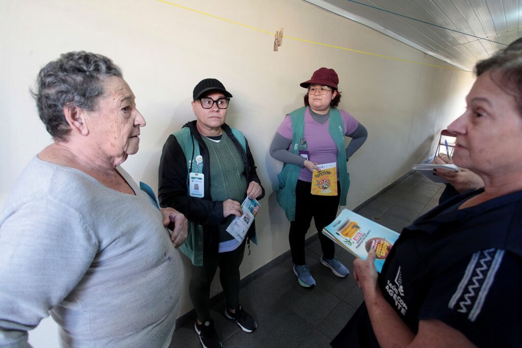 Esta fotografia registra um momento de interação social e profissional, onde profissionais da saúde comunitária dialogam com uma senhora em um ambiente interno, possivelmente durante uma campanha informativa ou visita domiciliar. A composição da cena é organizada de forma linear ao longo de um corredor estreito, com os sujeitos dispostos em um arranjo que sugere uma conversa em andamento. À esquerda, uma senhora está de perfil, voltada para uma profissional que está à direita, criando um eixo de comunicação principal. As outras duas profissionais, posicionadas centralmente, observam a interação, estabelecendo um ritmo visual que conduz o olhar do espectador pelo espaço. O enquadramento é em plano médio, capturando as expressões faciais e o contexto da atividade sem isolar os indivíduos do ambiente comum. Os sujeitos centrais são quatro mulheres. À esquerda, uma senhora de cabelos grisalhos curtos veste uma blusa de tons claros, mantendo uma postura de escuta. No centro, duas profissionais usam coletes verdes sobre roupas casuais, uma com boné e óculos, e a outra com um boné bordô, segurando materiais informativos. À direita, vemos a parte posterior e o perfil de uma quarta mulher, vestindo um uniforme escuro com a identificação "Agente de Saúde", que segura um folheto e parece estar conduzindo o atendimento. Todas apresentam uma postura séria e atenta, característica de um trabalho de assistência social. A imagem é uma fotografia documental digital, com um estilo realista e direto, típico de registros de campo ou reportagens locais. A execução técnica privilegia a nitidez nas expressões faciais e nos detalhes dos vestuários profissionais, com um foco mantido nos personagens principais. O estilo é desprovido de manipulações artísticas, focando na clareza da mensagem e na representação fiel da interação humana em um contexto público ou institucional. A iluminação é suave e uniforme, típica de ambientes internos, garantindo que não haja sombras fortes que distraiam o espectador. O cenário é um corredor de uma construção simples com paredes de cor clara, que confere uma sensação de sobriedade e neutralidade ao ambiente. O piso de cerâmica e a presença de um varal ao fundo indicam um espaço funcional, possivelmente um centro comunitário ou uma residência. A iluminação é difusa, vinda de fontes externas ao enquadramento, o que suaviza os contornos e confere um tom cotidiano à cena. A atmosfera geral é de seriedade e engajamento profissional, refletindo a rotina de um atendimento de saúde comunitária voltado ao bem-estar da população local.