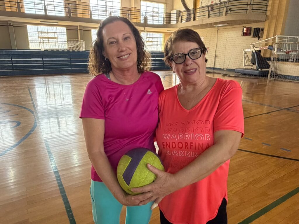 Duas mulheres posam juntas no centro de uma quadra poliesportiva coberta. Ambas sorriem para a câmera e seguram uma bola nas mãos compartilhadas. Pessoas e expressão Mulher à esquerda: cabelos castanhos ondulados na altura dos ombros, pele clara, veste camiseta rosa choque Adidas e calça esportiva azul claro. Sorriso leve e postura relaxada. Mulher à direita: cabelos curtos castanhos, usa óculos de grau e camiseta rosa/laranja com a palavra “ENDORFINA” impressa; também sorri e está mais próxima da câmera. As duas transmitem amizade, cumplicidade e energia positiva; não posso identificar quem são. Ação e objeto Ambas seguram uma bola amarelo‑verde com faixa roxa, como se a estivessem apresentando ou passando entre si.