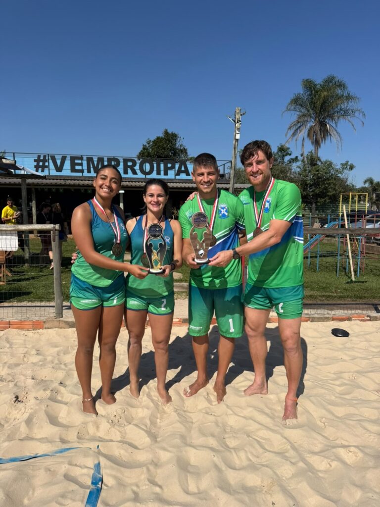 Foto de quatro atletas em quadra de areia posando com troféus e medalhas. Pessoas: quatro competidores alinhados (duas mulheres à esquerda, dois homens à direita), sorrindo e com os braços ao redor uns dos outros. Roupa: uniformes esportivos verdes com detalhes em azul e branco; números visíveis nas bermudas (2 e 1). Premiação: cada um usa medalha no pescoço; as duas pessoas do centro seguram troféus ornamentados. Local: quadra de areia (provavelmente vôlei de praia ou futevôlei) com linha azul no piso de areia. Fundo: estrutura com letreiro "#VEMPROIPA" atrás, cercado por grades; área com grama, árvores e um coqueiro ao fundo; céu azul claro e ensolarado. Luz e clima: dia claro, iluminação natural forte, sombras nítidas na areia. Atmosfera: momento de comemoração e vitória em evento esportivo ao ar livre.