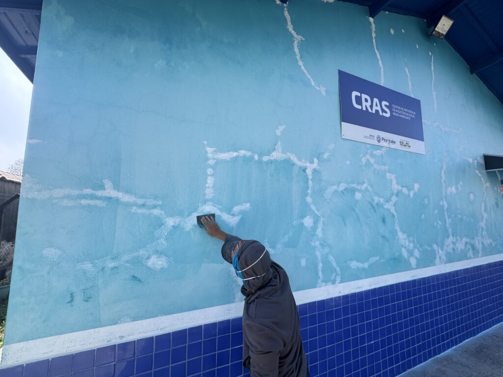 Parede externa azul em processo de reparo, com uma pessoa trabalhando em primeiro plano — vista de perfil e de costas, sem identificação possível. Composição: grande área da parede ocupa a maior parte da imagem; a pessoa está no terço inferior-direito, estendendo o braço para alisar/raspar a superfície com uma esponja ou lixa. Ação: mão segurando ferramenta de mão (possivelmente bloco de lixa ou esponja abrasiva) aplicando movimentos circulares/longos sobre a parede — clara atividade de preparo ou retoque do reboco/pintura. Superfície: parede pintada em tons de azul turquesa com muitas áreas de massa ou rejuntamento branco espalhadas (linhas e manchas irregulares), indicando correções de fissuras ou infiltrações antes da nova pintura. Faixa inferior: fileira de azulejos cerâmicos quadrados azul-escuro na base, com faixa branca entre azulejos e a parte pintada acima. Detalhes arquitetônicos: beiral/soalho de teto azul escuro visível no topo esquerdo; luminária externa no canto superior direito; pequena estrutura (janela ou grade) à direita parcialmente visível. Iluminação e clima: luz natural, céu claro ao lado esquerdo; sombras suaves que indicam dia ensolarado. Placa institucional: placa retangular azul afixada à parede à direita com letras grandes “CRAS” e texto menor (indica Centro de Referência de Assistência Social), confirmando uso público/institucional do local.