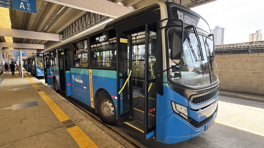 Um terminal de ônibus coberto com vários coletivos alinhados; em destaque, um ônibus azul e turquesa estacionado com a porta dianteira aberta no lado direito da imagem. Primeiro plano: frente e porta do ônibus à direita — degraus visíveis, barras e corrimões amarelos, adesivo de cadeira de rodas no para-brisa. Rodas e parte da lateral do ônibus com faixas coloridas (azul/turquesa) e o selo da Prefeitura de Piracicaba. Meio da imagem: mais ônibus encostados na mesma plataforma formando uma fila; letreiros eletrônicos nas laterais indicando números/rotas (por exemplo "123" e "508 SANTA RITA" parcialmente legível). Plataforma: pavimento de concreto com pisos táteis amarelos e faixas antiderrapantes; colunas e vigas do teto formando repetição visual até o fundo. Placas azuis de sinalização indicando posições de embarque (A02, A03 etc.). Fundo e ambiente: poucas pessoas aguardando ou caminhando pela plataforma à esquerda; parede de blocos à direita; prédios altos visíveis além do terminal. Iluminação natural forte, céu claro, sombra suave sob a cobertura. Impressão geral: cena típica de um terminal urbano em atenção ao embarque, organizada, com cores vivas nos ônibus e sinalização clara para passageiros.