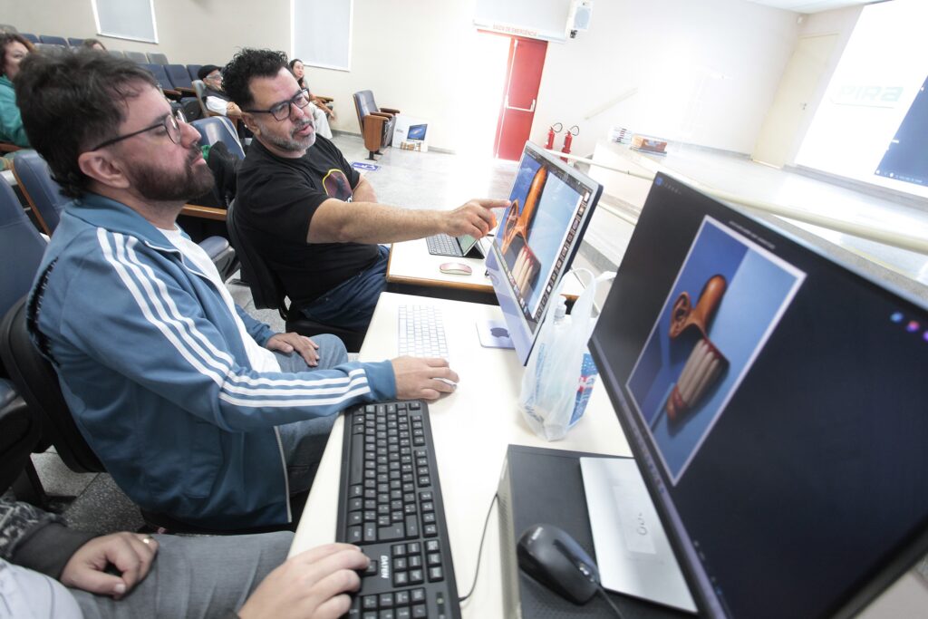Foto de uma sala tipo auditório onde duas pessoas estão trabalhando juntas em computadores de mesa. Cena principal: dois homens sentados lado a lado em cadeiras, voltados para monitores iMac colocados sobre uma mesa. O homem à direita (com camiseta preta) aponta para a tela do computador, orientando o colega; o homem à esquerda (com jaqueta azul claro esportiva) observa atentamente e controla o teclado/mouse. Computadores e conteúdo: telas mostram imagens em edição/visualização — uma delas exibe uma ilustração 3D ou manipulação digital de um objeto longo e curvo (tom alaranjado), outra mostra um close de uma orelha estilizada. Há teclado e mouse visíveis em primeiro plano. Ambiente: auditório com fileiras de poltronas ao fundo e algumas pessoas sentadas. Ao fundo, porta vermelha de emergência e extintores; iluminação predominante é natural/forte, com muita luz vindo da direita. Atmosfera: interação de ensino/colaboração técnica — parece uma oficina, curso ou sessão prática de design/edição digital. Expressões concentradas e postura colaborativa. Detalhes extras: objetos pessoais como sacola plástica e máscara cirúrgica sobre a mesa; piso claro e parede branca, sugerindo espaço institucional ou educacional.