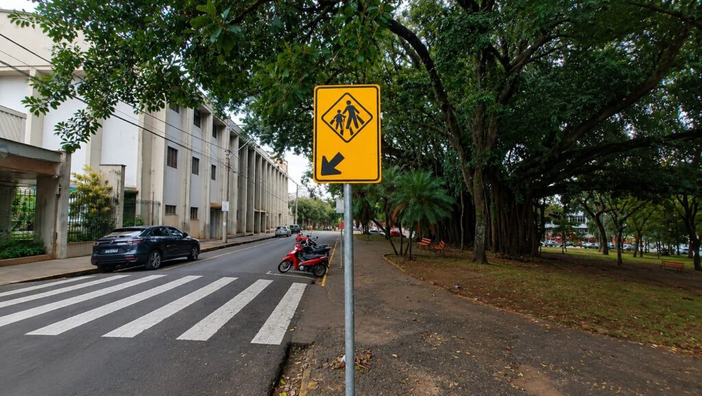 Foto de uma rua urbana com faixa de pedestres e sinalização indicando passagem de pedestres à esquerda. Elemento central: placa amarela com pictograma de adulto e criança dentro de um losango e uma seta preta apontando para baixo-esquerda — sinal alerta para travessia/ponto de pedestres. Faixa de pedestres: pintura em zebrinha branca no asfalto, em primeiro plano, atravessando a pista. Lado esquerdo: calçada e prédios de fachada clara (aparente prédio institucional ou comercial); um carro escuro estacionado junto à guia; postes e fiação aérea. Lado direito: parque/área arborizada com grandes árvores (copas densas), bancos laranjas e caminho de terra/areia; motos estacionadas próximas ao meio-fio. Trânsito e clima: via sem fluxo intenso (poucos veículos visíveis); iluminação difusa indicando dia nublado ou sombra forte das árvores. Ambiência: local parece calmo e arborizado, com infraestrutura para pedestres e área verde contígua à rua.