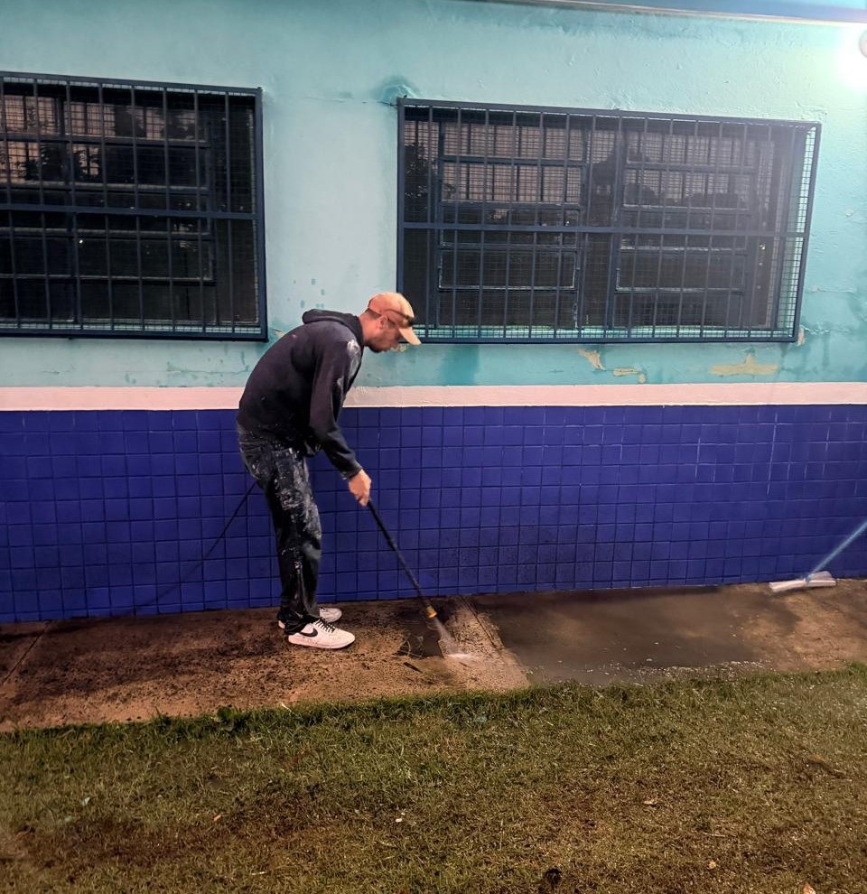 Homem limpando o piso ao lado de um prédio, visto de perfil. Pessoa: um homem vestido com moletom escuro, calça escura com respingos de tinta e tênis brancos, usando boné bege. Não faço identificação. Ação: ele segura uma mangueira/lança de jato (parece lavagem com água de alta pressão) e está fazendo a limpeza do passeio de concreto junto à parede. Parede e janelas: fachada pintada em tons de turquesa/verde claro na parte superior e azulejos quadrados azuis na metade inferior; duas janelas com grades metálicas pretas. Piso e área: faixa de concreto molhada onde ocorre a limpeza; faixa de grama verde em primeiro plano separando o passeio da câmera. Iluminação e clima: cena provavelmente ao entardecer/noite com iluminação artificial forte vindo da direita (lampião), criando sombras e reflexos no piso molhado.