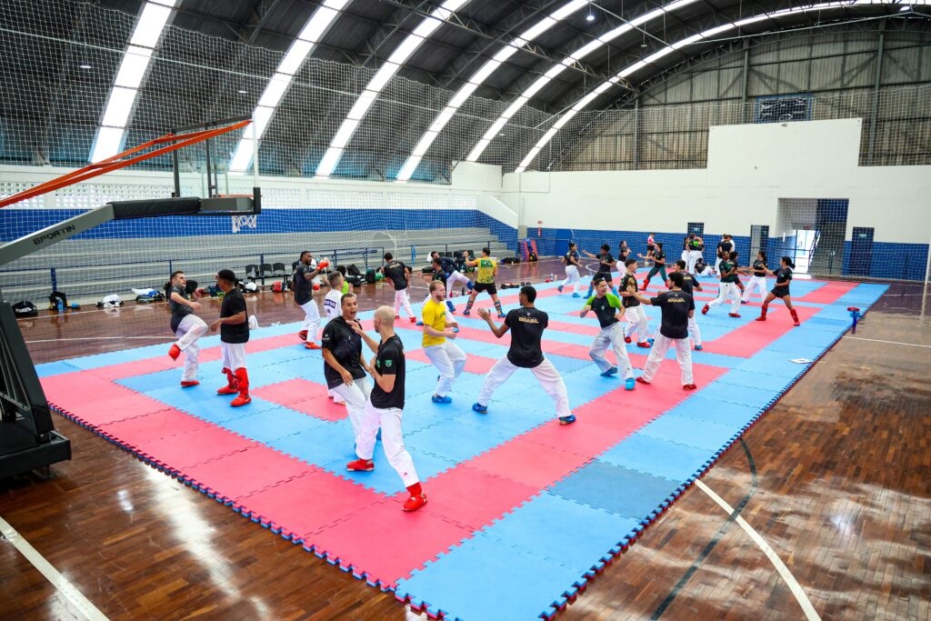 Esta é uma imagem que mostra um grupo de atletas praticando caratê (ou uma arte marcial similar) em uma quadra poliesportiva coberta. Ambiente: A quadra possui um piso de madeira, sobre o qual foram montados tapetes de EVA (tatames) nas cores azul e rosa, criando uma grande área de treinamento. O teto é alto e arqueado com estrutura metálica. Atletas: Vários praticantes, homens e mulheres, estão espalhados pela área. Eles vestem roupas esportivas: muitos usam camisetas pretas ou coloridas, calças de quimono brancas e proteções de canela e pés (caneleiras) na cor vermelha. Eles estão em duplas ou pequenos grupos, realizando movimentos de luta, socos e chutes, simulando um treino de combate (kumite). Estrutura: Pode-se ver uma tabela de basquete à esquerda e redes de proteção nas laterais da quadra. O espaço parece ser um ginásio bem iluminado e adequado para a prática esportiva.