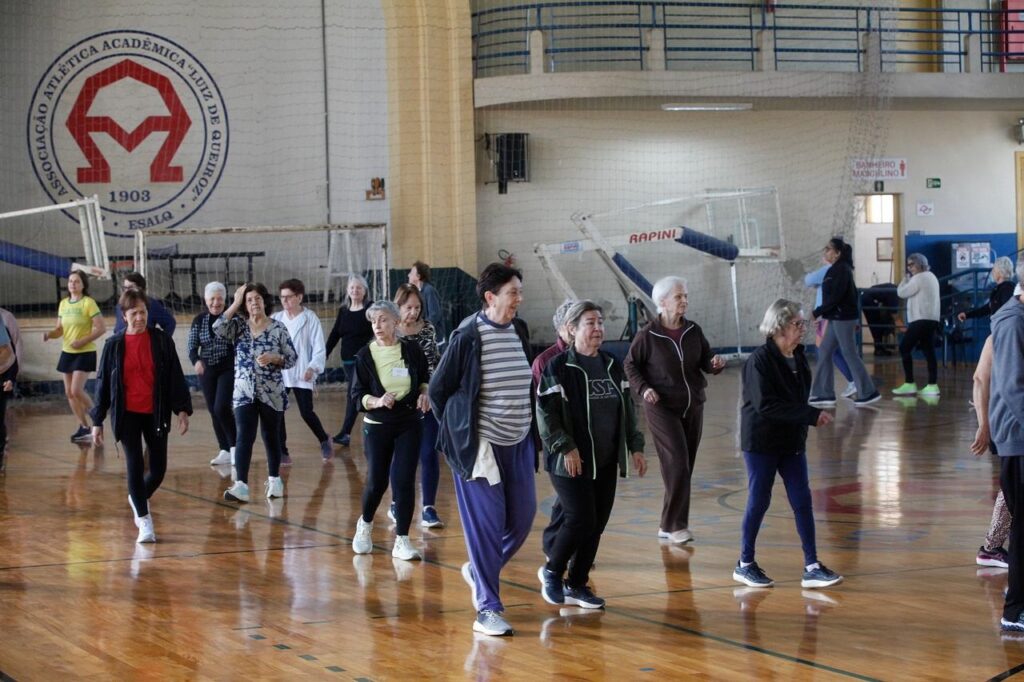 Grupo de pessoas fazendo atividade física em uma quadra poliesportiva coberta. A maioria caminha em formação, como num exercício coletivo ou aula de caminhada/gym para idosos. Piso de madeira polida com marcações esportivas; redes e gol ao fundo; logo grande na parede à esquerda. Pessoas e ação Predominância de pessoas idosas (mulheres) — roupas esportivas confortáveis: calças, agasalhos, camisetas e tênis. Movimento homogêneo: passos sincronizados, braços levemente movimentos; expressão concentrada e descontraída. Algumas pessoas ao fundo conversam ou aguardam, outras seguem o mesmo exercício. Ambiente e detalhes Quadra ampla, bem iluminada por luz natural; arquibancada superior e corrimões visíveis. Placas e equipamentos esportivos (rede, estrutura de basquete desmontada) na parede. Marcação de segurança e sinalização de banheiro ao fundo; atmosfera de atividade comunitária organizada.