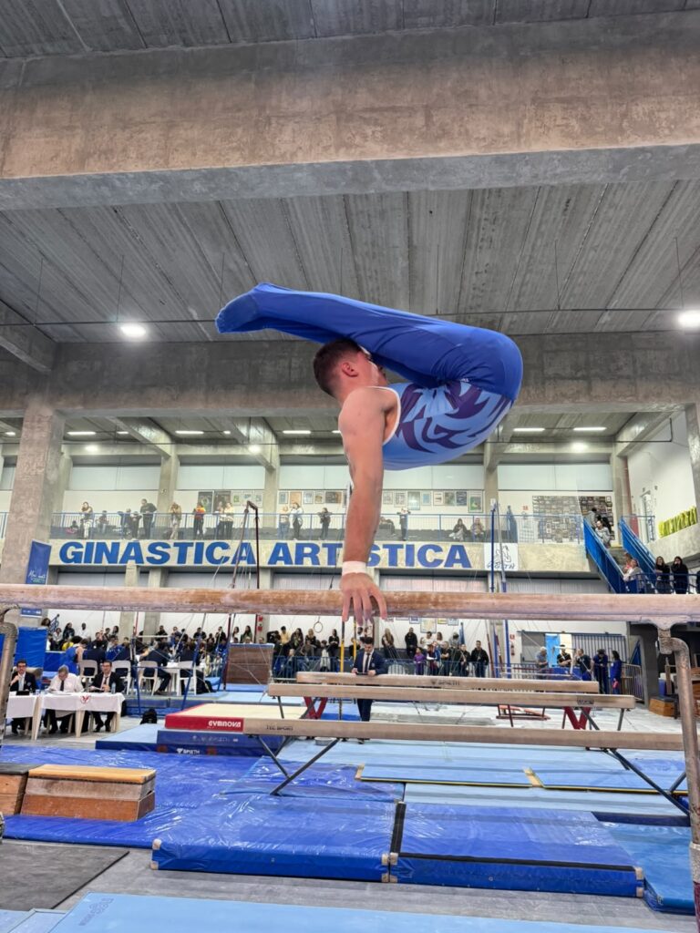 Nesta foto, um ginasta masculino está realizando um movimento impressionante nas barras paralelas em um ginásio de esportes. Aqui estão os detalhes: A Ação: O atleta está em uma posição de parada de mão, mas com o corpo dobrado em um ângulo agudo (em formato de "V" ou pique), mantendo o equilíbrio sobre as barras. Ele usa uma malha azul com detalhes em tons mais claros e calças longas azuis. O Ginásio: O ambiente é um centro de treinamento de "Ginástica Artística", como indica o letreiro bem visível ao fundo. O Contexto: Ao fundo, é possível ver arquibancadas com espectadores assistindo ao treino ou competição, além de outros equipamentos de ginástica espalhados pelo espaço. O clima parece ser de foco total na performance esportiva.