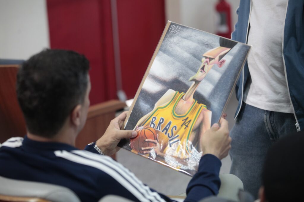 Esta foto mostra duas pessoas manuseando um quadro ou pôster com ilustração de um jogador de basquete. Ação: uma pessoa sentada (vista de costas) segura o quadro com as duas mãos; outra pessoa em pé segura a parte superior do quadro. Objeto: ilustração de um jogador magro e alongado, com regata amarela escrita "BRAS" e número 14, segurando uma bola de basquete; estilo caricatural e cores vibrantes. Ambiente: ambiente interno, possivelmente uma sala de reuniões ou auditório (cadeiras visíveis no canto inferior); fundo com portas ou painéis vermelhos desfocados. Detalhes: a pessoa sentada usa agasalho azul com listras brancas; a pessoa em pé veste camiseta branca e jaqueta azul. O quadro está protegido por plástico brilhante que reflete a luz.