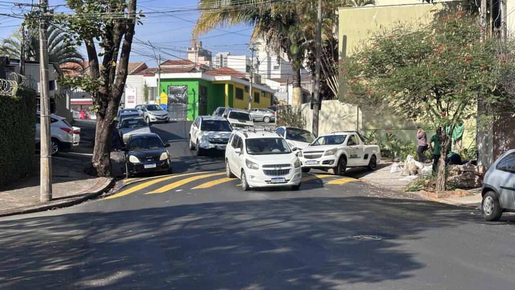 Cena diurna urbana em uma rua inclinada com tráfego e atividade de jardinagem no lado direito. Rua: inclinação suave para cima, faixas amarelas pintadas no asfalto em ondulação (marcação de lombada/atenção) no primeiro plano; vários carros circulando e estacionados em ambos os lados, predominando veículos brancos. Tráfego: um carro branco em destaque vindo em direção à câmera; outros carros sobem e descem a ladeira, formando um fluxo contínuo. Edificações: ao fundo, uma pequena construção com fachada pintada em verde e amarelo ocupa a esquina; mais ao fundo aparecem telhados de casas e alguns edifícios altos no horizonte. Vegetação e calçadas: árvores grandes com sombra projetada sobre a pista; à direita, um arbusto florido com flores vermelhas perto de um muro amarelo. Pessoas/atividades: do lado direito, três a quatro pessoas uniformizadas (camisas verdes) trabalhando na calçada, manuseando sacos e material de jardinagem/terra; outra pessoa com roupa estampada (rosa) caminha próxima. Infraestrutura: poste de madeira com muitos cabos elétricos e telefonia cruzando a cena; muros com cercas e arbustos à esquerda; calçadas estreitas com leve desnível. Iluminação/clima: dia claro e ensolarado, sombras bem definidas indicando luz forte; atmosfera típica de rua residencial/comercial movimentada.