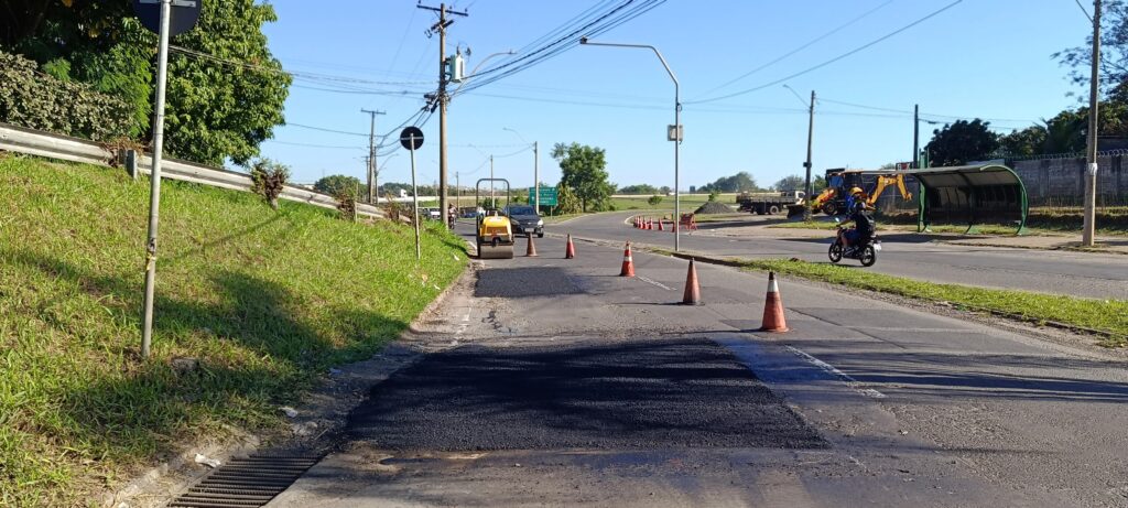 Uma cena diurna de reparo viário numa via urbana ampla, vista em formato panorâmico. No primeiro plano há remendos de asfalto recém-aplicados (áreas escuras) alinhados ao lado direito da pista, com cones laranja posicionados marcando a faixa de trabalho. Mais à frente há um rolo compactador amarelo parado na pista; ao fundo, um caminhão e uma retroescavadeira indicam operação em andamento. Dois motociclistas (um mais próximo, à direita) e alguns carros circulam pela via, desviando da área de serviço. À esquerda há um talude gramado com guard-rail e postes de sinalização; canteiro central baixo com grama separa as faixas. Do lado direito há uma estrutura de abrigo de ônibus verde e um muro com cercas; fios e postes elétricos cruzam o céu claro, sem nuvens. A iluminação é forte e gera sombras definidas, sugerindo manhã ou fim de tarde; a cena transmite trabalho prático de manutenção de pista em um dia ensolarado.