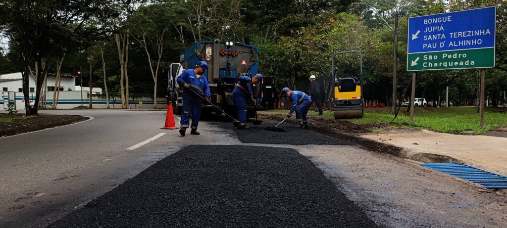 Foto de trabalhadores fazendo recapeamento/remoção e reposição de asfalto em uma via urbana. Ação principal: três operários com macacões azuis e capacetes mexem e espalham asfalto quente; um caminhão basculante de pavimentação está parado logo atrás deles despejando o material; um rolo compactador amarelo está à direita, pronto para compactar. Posição e enquadramento: tomada baixa e centralizada na faixa recém-asfaltada que domina a parte inferior da imagem, conduzindo o olhar até os trabalhadores ao centro. Sinalização e segurança: cone laranja próximo ao caminhão sinaliza a obra; os trabalhadores usam EPIs (capacetes e roupas de segurança). Ambiente: via curva ladeada por árvore e canteiro gramado; ao fundo há uma construção branca com grade e muitas árvores — parece uma área arborizada ou condomínio institucional. Placa de trânsito: painel grande à direita indica destinos (Bongué, Jupiá, Santa Terezinha, Pau d’Alhinho; setas para São Pedro e Charqueada). Detalhes do trecho: sarjeta e gradinha azul de drenagem no canto direito; solo ainda úmido e asfalto novo com cor escura contrastando com o pavimento antigo.