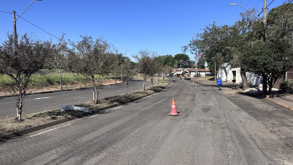Na imagem, observa-se um trecho urbano passando por manutenção viária, onde está sendo realizado o serviço de fresagem do pavimento. O asfalto antigo foi removido por meio de máquinas específicas, deixando a superfície com textura áspera e pronta para receber uma nova camada de revestimento. Cones de sinalização delimitam a área de trabalho, enquanto trabalhadores em uniforme azul operam os equipamentos, como o rolo compactador e a fresadora, garantindo que a preparação seja feita de forma adequada para melhorar a qualidade e durabilidade da via. Esse processo é essencial para assegurar um trânsito mais seguro e confortável após a repavimentação.