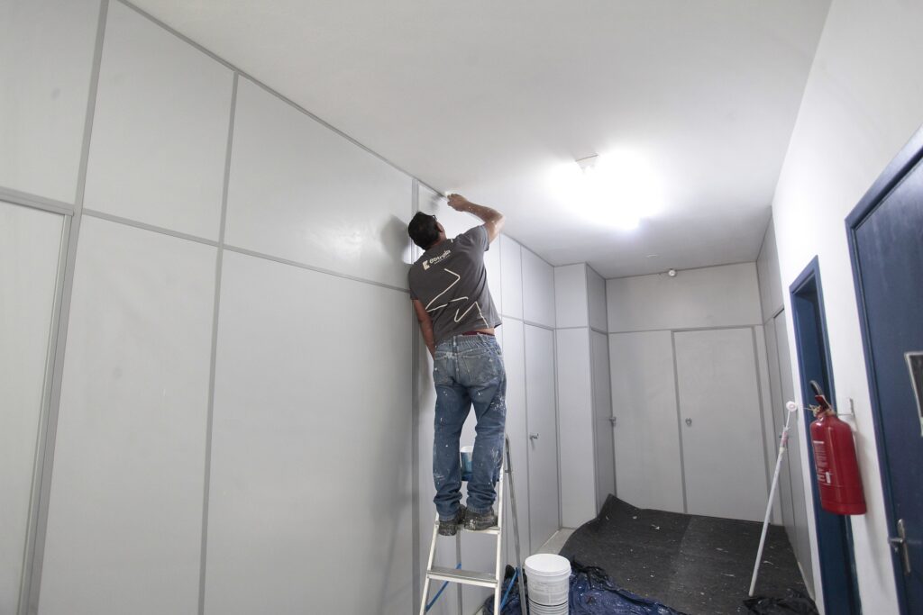 Nesta imagem, vemos um trabalhador fazendo o acabamento ou retoques de pintura em um ambiente interno. Aqui estão os detalhes: O trabalhador: Um homem está em cima de uma escada dobrável, usando uma camiseta cinza (com o logo da empresa "estrela" nas costas) e calça jeans. Ele está de costas, aplicando tinta branca cuidadosamente no topo da parede, perto do teto. O local: É um corredor ou sala com paredes formadas por painéis modulares cinza-claros, que dão um ar profissional e clean ao ambiente. Há várias portas lisas no fundo. Equipamento e organização: No chão, há uma lona preta protegendo o piso contra respingos. Perto da escada, vemos um balde de tinta e um rolo de pintura encostado na parede. Detalhe: À direita, fixado na parede próximo a uma porta com batente azul, há um extintor de incêndio vermelho, completando o cenário de um ambiente comercial ou institucional em manutenção.