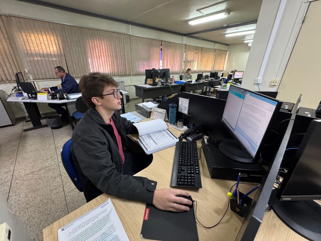 Um jovem em um escritório com vários colegas trabalhando em computadores. A imagem retrata um ambiente de escritório movimentado, com vários funcionários trabalhando em seus computadores. No primeiro plano, um jovem com óculos usa um laptop e segura um mouse, concentrado em sua tarefa. Ele veste uma jaqueta escura sobre uma camiseta rosa e tem um caderno aberto à sua frente, com papéis espalhados em sua mesa. Ao lado dele, há um teclado, um telefone, uma garrafa de água e um pequeno calendário. Ao fundo, outros colegas podem ser vistos em suas estações de trabalho, com monitores de computador e pilhas de documentos. A iluminação é artificial, proveniente de lâmpadas no teto, e a luz natural entra pelas janelas ao fundo, filtrada por persianas, criando um ambiente de trabalho típico, com uma atmosfera produtiva e focada. O chão é de granilite e as paredes são claras, contribuindo para a funcionalidade do espaço.