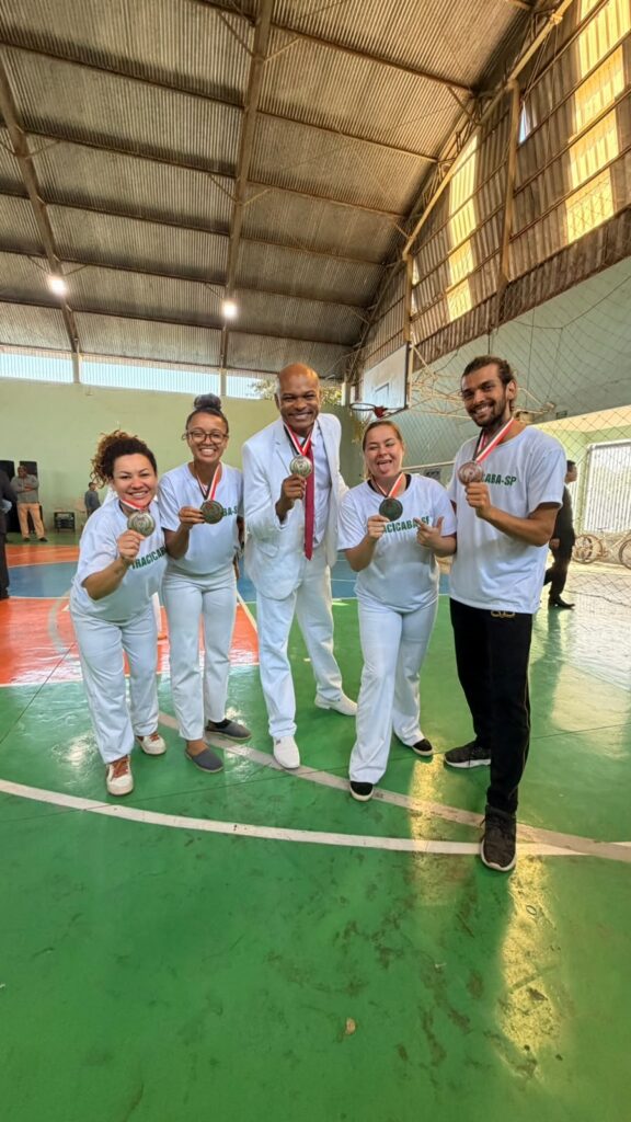 Grupo de cinco pessoas posando em quadra coberta, todas sorrindo e mostrando medalhas penduradas no pescoço. Composição: cinco pessoas alinhadas de frente para a câmera; a figura central é um homem vestido com terno branco e gravata vermelha, os outros quatro vestem camisetas brancas com inscrição verde (duas mulheres à esquerda, uma mulher e um homem à direita). Medalhas: todos seguram e exibem medalhas prateadas com fita vermelha e branca. Roupas e calçados: predominância de branco nas roupas (calças e camisetas), calçados variados — tênis e sapatos fechados; o homem à direita usa calça preta. Ambiente: quadra esportiva interna com piso pintado em verde, laranja e azul; rede e tabela de basquete ao fundo; estrutura de telhado metalizada e janelas altas deixando entrar luz. Iluminação e clima: luz natural e artificial misturadas, clima festivo e de comemoração após competição ou evento esportivo.