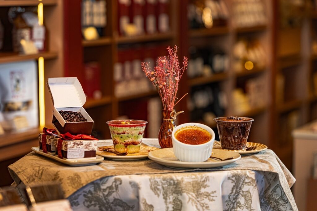 Esta imagem mostra uma mesa delicadamente arrumada com uma seleção de sobremesas gourmet, ambientada em um local que parece ser uma doceria ou loja de chocolates fina. Aqui estão os detalhes do que está sendo servido: À esquerda: Uma caixa de presente aberta, contendo doces (provavelmente brigadeiros cobertos com granulado), sobre outra caixa menor decorada com laço vermelho e um padrão clássico. Ao centro (esquerda): Um copinho plástico transparente com uma sobremesa em camadas: base de massa ou creme, uma camada verde (possivelmente pistache), um recheio escuro e uma cobertura avermelhada ou de frutas. Decoração central: Um pequeno vaso de vidro âmbar com um ramo de flores secas rosadas/avermelhadas, adicionando um toque de elegância ao centro da mesa. À direita: Um ramequim branco contendo um crème brûlée, com aquela crosta caramelizada característica no topo. Ao lado dele, outro copinho transparente contendo uma sobremesa de chocolate (como uma mousse ou ganache). Contexto: Tudo está disposto sobre uma mesa coberta com uma toalha de tecido estampado em tons claros e cinza. Ao fundo, vemos estantes desfocadas contendo mais produtos, o que sugere um ambiente comercial aconchegante e sofisticado. A iluminação é quente e focada nas sobremesas.