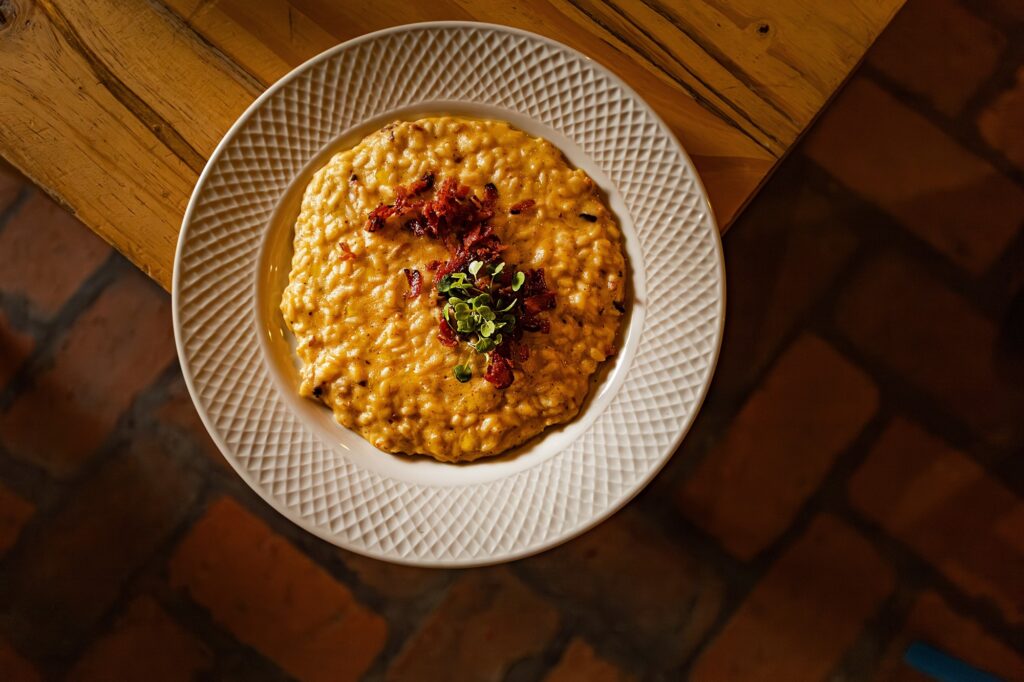 Prato visto de cima: risoto cremoso servido num prato branco com borda texturizada, posicionado levemente deslocado para a esquerda sobre uma mesa de madeira rústica. Detalhes observáveis: Aparência do prato: risoto de cor amarelada/dourada, textura cremosa e brilhante com arroz bem envolvido no molho. Guarnecimento: lascas crocantes escuras por cima (aparentam ser bacon ou prosciutto crocante) e um tufo de microverdes/ervinhas no centro, trazendo cor verde viva. Prato e superfície: prato branco com padrão geométrico na borda; mesa de madeira clara com grãos e ranhuras visíveis à esquerda. Ao fundo, piso de cerâmica avermelhada desfocado. Iluminação e cor: luz quente e direcionada, realçando brilho do molho e tons dourados do arroz; sombras suaves criam sensação acolhedora. Sensação e contexto: imagem transmite um prato robusto e reconfortante, provável entrada principal ou prato principal em restaurante casual/acolhedor; apresentação simples porém apetitosa.