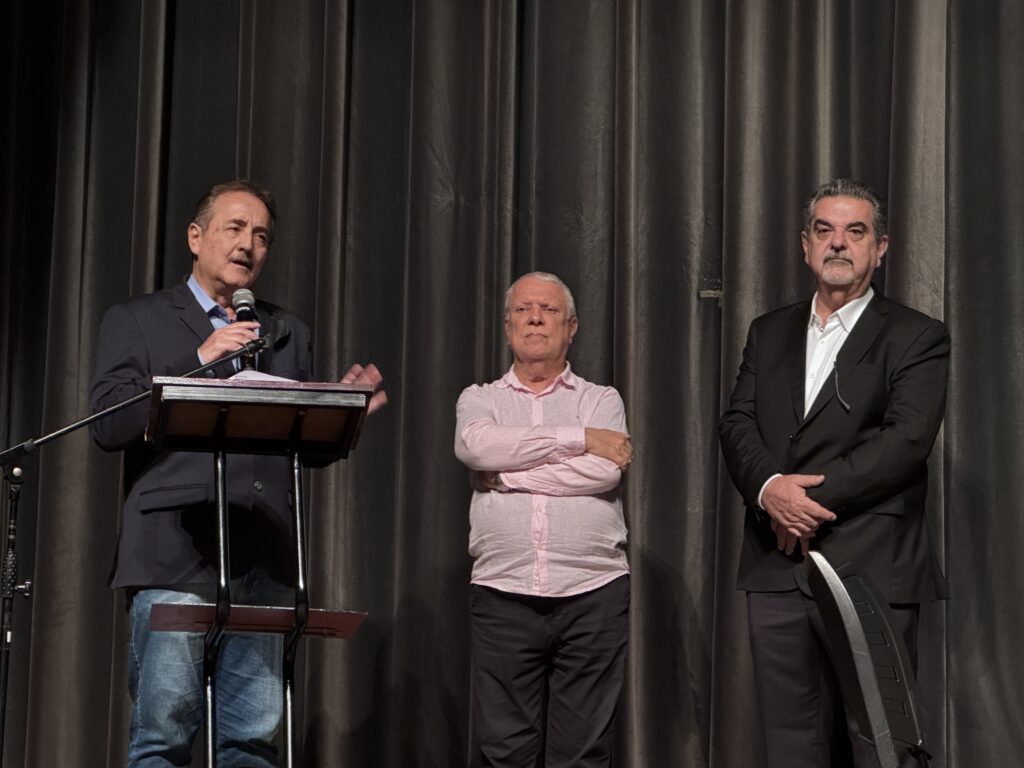 Nesta foto, três homens estão em um palco diante de uma cortina escura. À esquerda, um homem de blazer azul marinho sobre uma camisa azul clara está em um púlpito, segurando um microfone e falando com um gesto na mão. Ele usa jeans. No centro, um homem usa uma camisa rosa de mangas compridas com os braços cruzados, olhando seriamente para frente. À direita, outro homem veste um terno preto com camisa branca por baixo, também com os braços cruzados e uma expressão serena. Parece ser um evento formal, como uma palestra ou cerimônia de premiação.