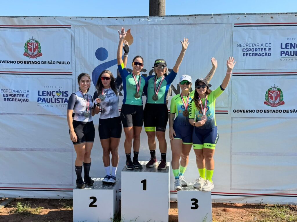 Foto de pódio ao ar livre com seis mulheres ciclistas comemorando suas colocações. Composição: três degraus de pódio numerados (1, 2, 3). Duas atletas ocupam o degrau do 1º lugar ao centro, duas no 2º lugar à esquerda e duas no 3º lugar à direita. Expressão e gesto: a maioria sorri e levanta os braços em comemoração; algumas seguram medalhas penduradas no pescoço. Roupa/equipamento: roupas de ciclismo justas (jerseys e bermudas); óculos esportivos, capacetes ou bonés, e meias altas visíveis em algumas atletas. As equipes têm cores distintas — uma com tons verde-azulado, outra com branco e preto, e outra com amarelo/neon e azul. Medais e troféus: medais de metal penduradas no pescoço de todas as competidoras; uma atleta do time do centro segura também um pequeno troféu. Cenário: painel de fundo com logos e textos institucionais (Governo do Estado de São Paulo, Secretaria de Esportes e Recreação, Prefeitura de Lençóis Paulista). Céu azul claro sem nuvens, indicando dia ensolarado. Piso e ambiente: solo de terra à frente do pódio, sombra leve; evento claramente esportivo, em área externa.