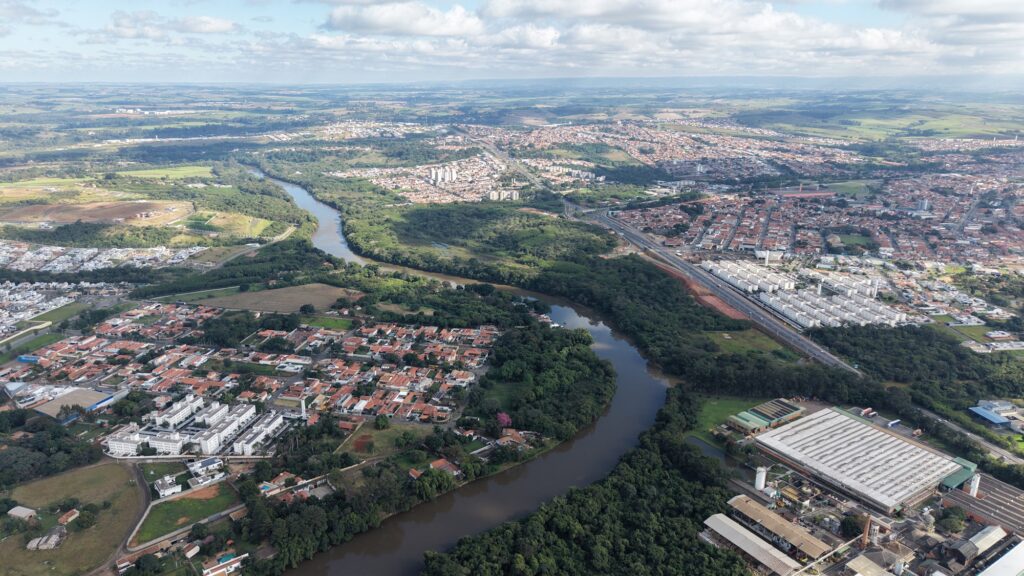 A imagem mostra uma vista aérea de uma cidade cortada por um rio sinuoso, que organiza e dá forma ao crescimento urbano. De um lado e de outro, espalham-se bairros residenciais com telhados vermelhos, áreas industriais e manchas de vegetação que suavizam o cenário. Ao fundo, surgem edifícios mais altos concentrados no centro, enquanto colinas onduladas delimitam o horizonte, criando um contraste marcante entre a expansão urbana e os elementos naturais que ainda resistem na paisagem.