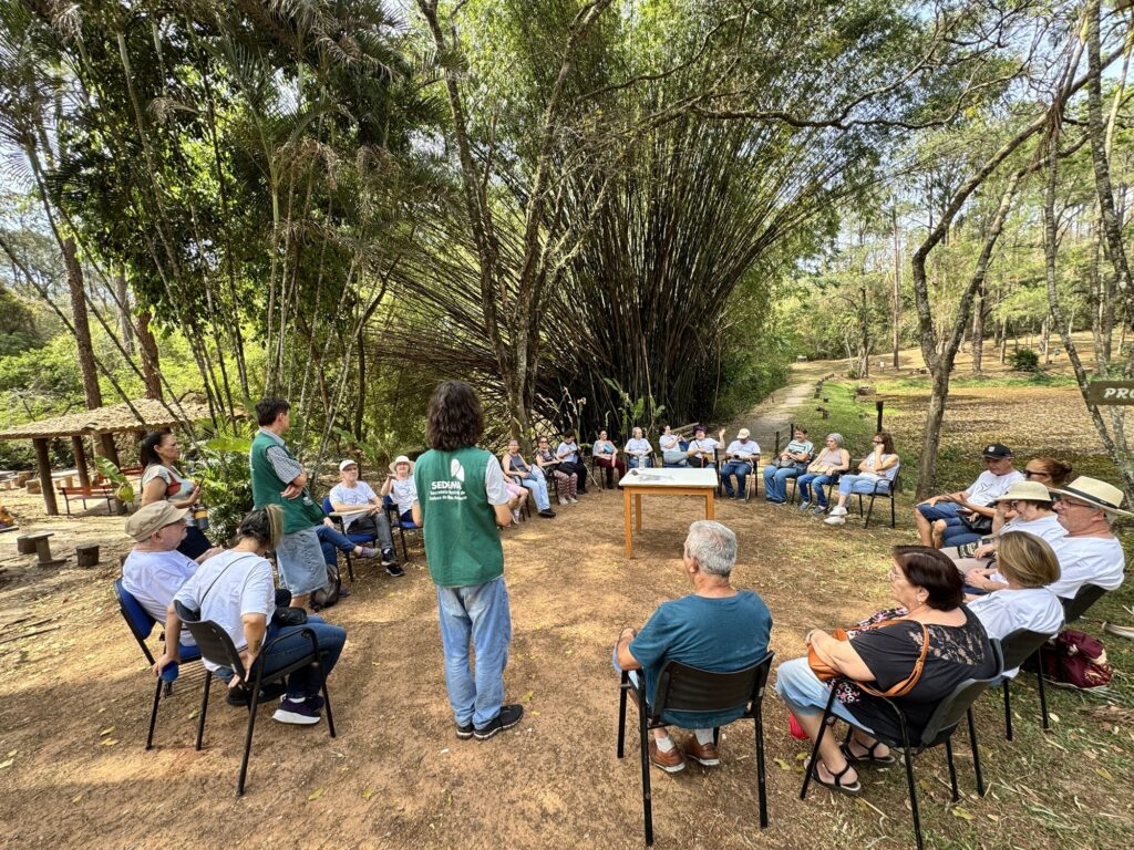 Esta imagem mostra um grupo de pessoas participando de uma atividade ao ar livre em um ambiente de parque ou reserva natural, sob a orientação de monitores. Aqui estão os detalhes principais: O Grupo: Há um grande círculo de cadeiras dispostas em um terreno de terra batida. As pessoas parecem ser, em sua maioria, idosos, sentados e atentos ao que está sendo apresentado. Os Monitores: No centro do círculo, há dois monitores (ambos vestindo coletes verdes da "SEDEMA - Secretaria Municipal de Defesa do Meio Ambiente"). Um deles está de costas para a câmera, aparentemente liderando a conversa ou atividade, enquanto o outro está de pé, à esquerda, observando o grupo. O Ambiente: O local é muito arborizado, com árvores altas ao fundo e, especificamente, um grande grupo de bambus que forma um "túnel" ou barreira natural ao fundo. Há também uma trilha ou caminho gramado que leva para dentro da mata à direita. Detalhes Adicionais: Há uma mesa de madeira vazia no centro do círculo, que parece servir como ponto de apoio. À esquerda, vê-se parte de um quiosque ou área coberta de palha. O clima parece ser de uma manhã ensolarada, com luz filtrada pelas árvores, criando uma atmosfera tranquila e educativa. O evento tem características de uma atividade de educação ambiental ou visita guiada para grupos da terceira idade.