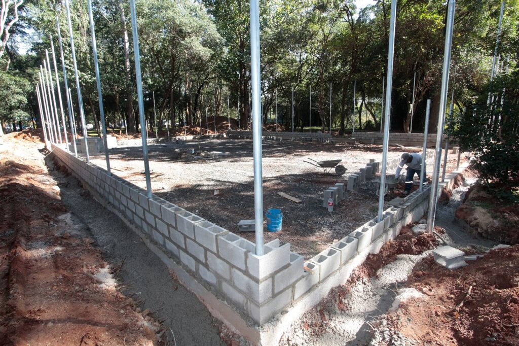 A imagem mostra uma obra em andamento em uma área aberta, com bastante vegetação ao redor. Aqui estão os detalhes principais: Estrutura: Está sendo levantado um muro baixo de blocos de concreto, que parece delimitar o perímetro de uma construção (possivelmente uma quadra, dado o formato e os postes). Postes: Há vários postes metálicos verticais fixados ao longo da estrutura de blocos, provavelmente para sustentar uma futura cerca ou tela de proteção. Trabalhador: Um operário está trabalhando no canto direito, usando um carrinho de mão para manusear materiais. Terreno: O interior da área delimitada é de terra batida/cascalho. Há materiais de construção espalhados, como blocos soltos, um balde azul e uma pá. Ambiente: A obra está localizada em um ambiente arborizado, com muitas árvores grandes e sombra ao fundo. O solo ao redor da construção parece ter sido recentemente escavado para a fundação.