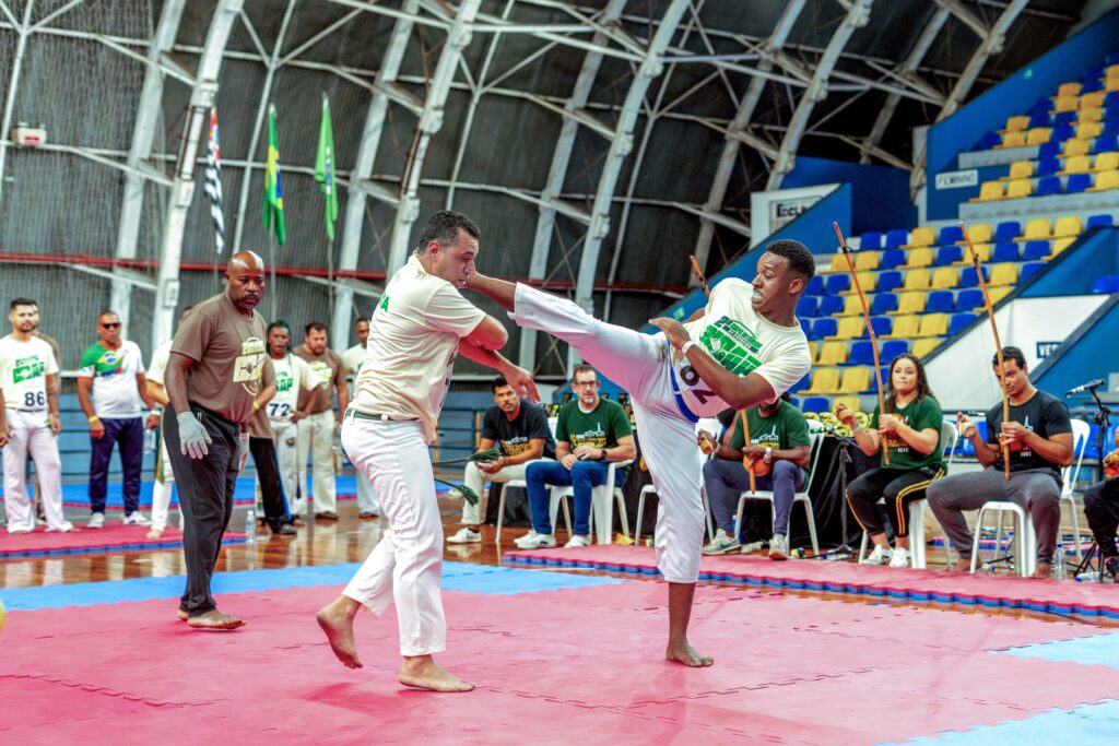 Esta imagem captura uma demonstração dinâmica de capoeira dentro de um ginásio esportivo. Ação Central: Dois homens estão no centro do tatame (composto por placas de espuma vermelhas e azuis). O capoeirista da direita está executando um chute alto (parecido com uma meia-lua), enquanto o capoeirista da esquerda se esquiva do golpe. Ambos vestem roupas típicas de capoeira: camisas brancas e calças brancas. Ambiente: O local parece ser um ginásio coberto com estrutura metálica aparente no teto. Ao fundo, há arquibancadas com assentos azuis e amarelos. Pessoas ao redor: Há várias outras pessoas observando a roda de capoeira. À esquerda, um homem de camisa marrom, que parece ser um instrutor ou árbitro, observa atentamente com as mãos na cintura. Mais atrás, outros participantes aguardam, alguns vestindo camisetas com logos de grupos de capoeira. À direita, sentados em cadeiras, estão os músicos da roda, segurando berimbaus e acompanhando o ritmo. Clima: A cena transmite muita energia, foco e tradição, típica de eventos de capoeira. As pessoas estão engajadas no movimento, que combina arte marcial, dança e cultura.