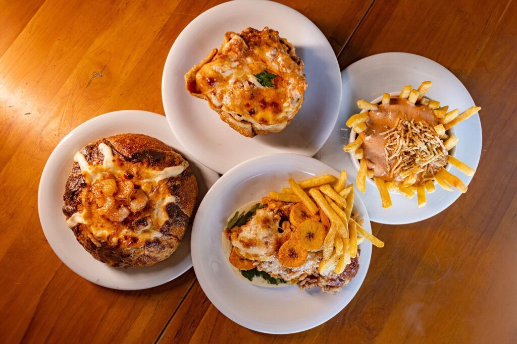 A foto mostra quatro pratos diferentes dispostos sobre uma mesa de madeira. Aqui está o que temos: No canto inferior esquerdo: Um pão tipo italiano recheado (parece ser um "escondidinho" ou creme de camarão), com o topo gratinado e camarões aparentes. No topo, ao centro: Um prato que parece ser uma torta ou lasanha individual, também bem gratinada e dourada. No canto inferior direito: Um prato com uma base que parece ser carne ou arroz coberto com queijo derretido, decorado com rodelas de banana frita e acompanhado de batatas fritas. À direita: Uma porção de batatas fritas servida em uma tigela, coberta com um molho cremoso (possivelmente molho rosé ou queijo) e batata palha por cima.