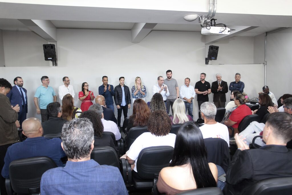 Esta imagem mostra um evento oficial ou uma palestra acontecendo em um auditório fechado. O palestrante: No centro do grupo de pessoas em pé, um homem de camisa azul clara está falando ao microfone, enquanto outras pessoas ao seu redor ouvem atentamente ou aplaudem. O público: Há uma plateia sentada em cadeiras pretas, de costas para a câmera, assistindo à apresentação. Algumas pessoas na plateia também estão aplaudindo. O grupo de apoio: Atrás das cadeiras, há uma linha de pessoas em pé, formando uma espécie de mesa de autoridades ou grupo de convidados, composto por homens e mulheres vestidos de forma social ou casual-chique. O ambiente: É uma sala com paredes neutras, iluminação interna, caixas de som instaladas nas paredes laterais e um projetor fixado no teto. O clima parece ser de uma reunião formal, possivelmente institucional ou política, dado o perfil do grupo e a organização do espaço.