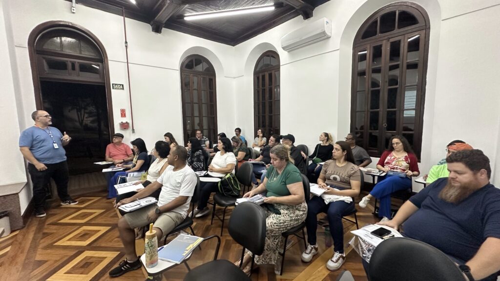 Nesta foto, vemos um grupo de pessoas participando de uma palestra ou treinamento em uma sala com arquitetura clássica. Aqui estão os detalhes principais: O Instrutor: À esquerda, um homem de camiseta azul está de pé, falando e gesticulando para o grupo. Ele parece ser o responsável pela apresentação. O Público: O grupo é composto por um público diverso, formado por adultos de várias idades, todos sentados em cadeiras dispostas em fileiras, atentos à explicação. Muitos deles têm materiais, cadernos ou apostilas sobre as pernas ou sobre pequenas mesas de apoio acopladas às cadeiras. O Local: A sala tem características marcantes, com piso de madeira em padrão geométrico, paredes brancas e janelas altas em formato de arco com esquadrias de madeira escura. Há um ar-condicionado na parede superior e um extintor de incêndio próximo à porta. O Clima: O ambiente transmite uma sensação de aprendizado e atenção, típica de um curso ou workshop.