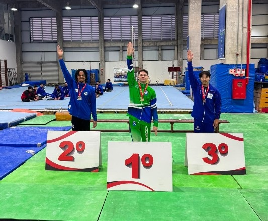 Esta foto mostra três atletas de ginástica no pódio, celebrando após uma competição em um ginásio. Aqui estão os detalhes: O Pódio: Os três estão posicionados atrás de placas que marcam as colocações (2º à esquerda, 1º ao centro e 3º à direita). Os Atletas: Todos usam agasalhos esportivos, medalhas no pescoço e estão com o braço direito levantado em um gesto de vitória. O atleta no 1º lugar (centro) veste um agasalho verde e azul. O atleta no 2º lugar (esquerda) usa uma jaqueta azul. O atleta no 3º lugar (direita) também veste uma jaqueta azul. O Cenário: Eles estão em um ginásio de ginástica artística, com tapetes azuis e verdes espalhados pelo chão e equipamentos de treino ao fundo, onde outras pessoas também estão sentadas.