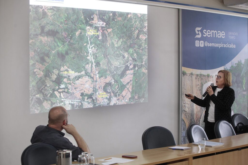 Esta foto registra um momento de apresentação profissional em uma sala de reuniões. Aqui estão os detalhes principais: A Apresentadora: Uma mulher de cabelos claros e óculos, vestindo um blazer escuro texturizado sobre uma blusa branca, segura um microfone e um controle remoto, enquanto aponta para o telão. O Telão: É exibido um mapa de satélite da região de Piracicaba. Nele, é possível identificar pontos como a "Usina Costa Pinto", "Vila Sônia", "Santa Teresinha" e a rodovia Charqueada, destacados com legendas. O Público: Em primeiro plano, vemos a parte de trás de um homem careca vestindo uma camisa cinza escura, que assiste à apresentação com atenção, apoiando o queixo na mão. O Local: A sala possui uma mesa de madeira longa, com copos de água e papéis. Ao fundo, há um banner vertical azul com a logomarca do "semae" (Serviço Municipal de Água e Esgoto de Piracicaba) e o slogan "UM NOVO TEMPO".
