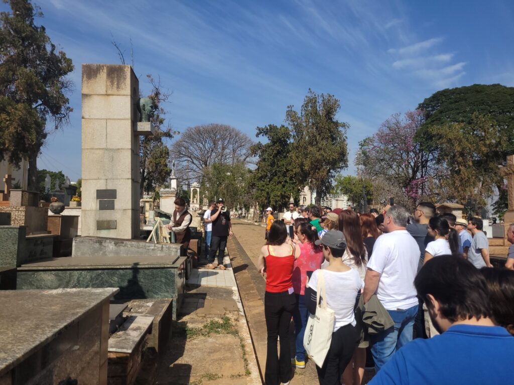 Foto de uma visita guiada/evento público num cemitério histórico, tirada em dia ensolarado. Observações detalhadas: Cena geral: grande grupo de pessoas reunidas ao lado direito da imagem, formando uma fila/aglomerado que olha na direção de um guia ou apresentação à esquerda. Público: diversidade de idades — jovens e adultos; muitos estão de pé, alguns registrando com celular; roupas casuais de primavera/verão (camisetas, regatas, bonés). Ninguém é identificado individualmente. Atividade central: um homem com megafone (de camiseta preta e boné) fala para o grupo; ao lado dele, outro homem vestido com traje de época/figurino (colete marrom, camisa clara) manipula suportes ou quadros sobre uma lápide, sugerindo encenação histórica ou demonstração. Túmulos e monumentos: vários jazigos e lápides de pedra/ mármore em primeiro plano e ao fundo; um monumento alto de blocos de pedra com uma escultura de rosto em bronze projetando-se lateralmente destaca-se à esquerda-central. Cruz e esculturas ornamentais aparecem à esquerda. Paisagem e arquitetura: via central calçada de terra/areia entre as alamedas de túmulos; árvores grandes ao fundo (algumas com poucas folhas) e vegetação; céu azul com nuvens finas, iluminação forte e sombras definidas — dia claro e seco. Composição e perspectiva: foto tomada atrás do público, olhando em direção ao guia e ao monumento; profundidade mostra caminho do cemitério e mais túmulos se estendendo ao fundo. Atmosfera: tom respeitoso, educativo e comunitário — aparenta ser passeio histórico, visita guiada ou reconstituição cultural relacionada ao patrimônio funerário. Detalhes observáveis: algumas pessoas usam sacolas/bolsas, uma mulher em primeiro plano veste regata vermelha; há pequenas áreas de grama seca ao lado do caminho; muitos monumentos têm placas e ornamentos clássicos.