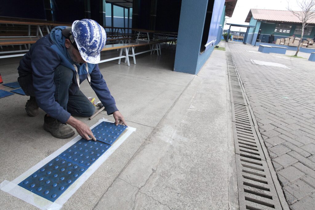 Homem agachado aplicando placas táteis azuis numa faixa de piso junto a um prédio — trabalho de acessibilidade em andamento. Pessoa: trabalhador visto de lado/trás, usando capacete azul manchado de tinta, moletom azul com capuz e calça jeans; está ajoelhado e concentrado no assentamento das placas. Ação: posiciona e pressiona painéis táteis azuis com relevo circular sobre uma fita branca já colocada no chão; há uma talhadeira/rodo e materiais próximos. Local: corredor coberto ou abrigo (possivelmente de escola ou prédio público) com longas mesas/banquinhos empilhados ao fundo; à direita abre-se pátio externo com piso intertravado e canaleta de drenagem. Ambiente e estado: área limpa e organizada, boxes/caixas empilhadas ao longe; iluminação natural difusa (céu nublado ou luz indireta). Sensação: cena de trabalho manual detalhado, foco em acessibilidade e manutenção do espaço público.