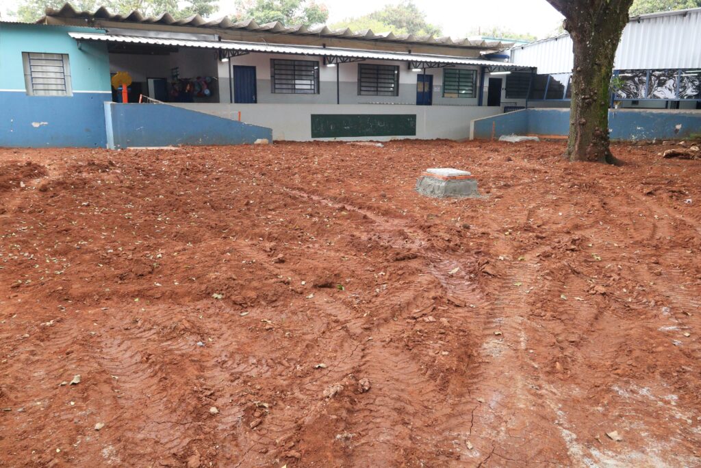 Campo de terra batida recém-removido em frente a um prédio de salas/creche/escola, vista panorâmica. Primeiro plano: grande área de solo vermelho revolvido, com pneus/roçadas visíveis e marcas de maquinário. Folhas secas espalhadas e sulcos irregulares indicam movimento recente de terra. Centro: bloco de concreto retangular levantado sobre uma base de argamassa, posicionado isoladamente sobre o solo — parece um marco, caixa de passagem ou preparação para poste/estrutura. Direita: árvore robusta com casca coberta de pequenos nódulos, enraizada na área de trabalho; parte do tronco aparece destacada contra o chão aberto. Fundo: prédio baixo com fachada pintada em tons de azul e cinza, com corrimões e pequenas rampas de acesso. Janelas com grades, portas azuis e um painel negro (possivelmente quadro ou lousa) fixado na parede. Cobertura: beiral com telhas e estrutura metálica sobre a faixa frontal do prédio, com iluminação instalada sob o beiral. Atmosfera/clima: cena diurna com iluminação difusa; ambiente sugere obra leve ou preparação de terreno para reforma/implantação de área externa, possivelmente em instituição escolar ou comunitária.