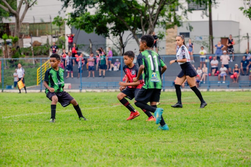 Nesta foto, vemos um jogo de futebol infantil acontecendo em um campo gramado ao ar livre. Em destaque, três meninos estão correndo durante a partida: O menino à esquerda veste uma camisa verde com detalhes pretos e calção preto. O menino no meio está com uma camisa vermelha e preta. O menino à direita, visto de costas, usa uma camisa verde e preta com o número 11 nas costas. Do lado direito, uma mulher, que parece ser a árbitra da partida, acompanha a jogada; ela usa um uniforme preto e branco. Ao fundo, atrás de uma grade azul, há várias pessoas assistindo ao jogo sentadas na arquibancada, e algumas árvores compõem a paisagem atrás do campo. O clima parece ser de uma tarde ensolarada e esportiva.
