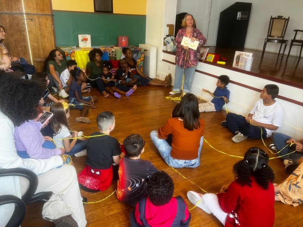 Uma cena encantadora de uma atividade de contação de histórias ou mediação de leitura. Um grupo diversificado de crianças e adultos está sentado em roda sobre um piso de madeira, em uma sala ampla. A atividade: Uma mulher adulta, em pé ao fundo, segura um livro aberto e conduz a dinâmica, que envolve um fio de barbante amarelo conectando as pessoas da roda, criando uma teia simbólica entre todos. Os participantes: Há várias crianças de diferentes idades e alguns adultos sentados no chão, todos prestando atenção. Alguns seguram o barbante, participando ativamente da interação. O ambiente: O local parece um centro cultural ou biblioteca, com um pequeno palco ao fundo onde outros livros estão expostos. Há também uma lousa verde na parede e cadeiras de madeira vazias no canto direito. Clima: A atmosfera é de aprendizado e conexão, com todos envolvidos na atividade lúdica e educativa.