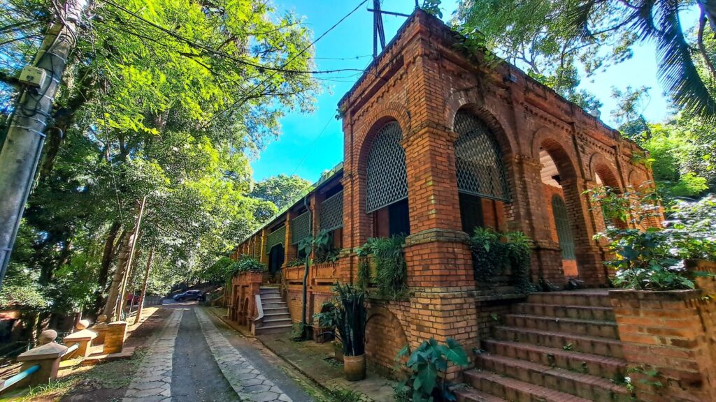 Um prédio antigo de tijolos à vista em estilo clássico-romano, visto em ângulo baixo à direita da foto. Principais detalhes: Arquitetura: alvenaria de tijolo vermelho com arcos grandes e colunatas retas; soleiras, cornijas e detalhes em relevo que sugerem construção histórica ou inspirada em arquitetura do século XIX/começo do XX. Fachada: janelas e vãos protegidos por grades em padrão treliça, entradas por degraus amplos em ambos os lados; alguns arcos estão abertos, outros fechados com portas ou grades. Vegetação: muitas árvores altas e folhagens ao redor — luz do sol atravessando as copas cria tons vibrantes de verde e sombras pontuais sobre o prédio e a rua. Rua e entorno: caminho estreito de paralelepípedos à esquerda, pequenas muretas e postes de iluminação; há um carro estacionado ao fundo, o que indica uso atual do local. Estado: bem conservado, com plantas ornamentais em floreiras e trepadeiras somando charme rústico; algumas áreas mostram musgo e sinais naturais de envelhecimento. Atmosfera: sensação tranquila, quase de refúgio urbano — mistura de historicidade com natureza exuberante em dia ensolarado.