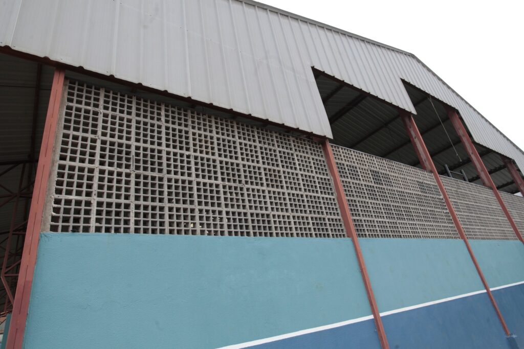 Vista inclinada de uma fachada/estrutura metálica e muro em ângulo baixo. Parte superior: telhado metálico corrugado claro sustentado por vigas e colunas metálicas pintadas de vermelho-oxidado; há aberturas retangulares sob o beiral que mostram o forro do telhado. Meia-altura: painel ventilado feito de blocos vazados (tijolos de ventilação) formando uma malha quadrada repetida, cobrindo grande extensão da parede. Parte inferior: parede lisa pintada em tons de azul claro e azul escuro, separadas por uma faixa branca horizontal; a pintura mostra pequenas irregularidades e marcas. Estrutura e materiais: combinação de aço, blocos de concreto e reboco pintado; algumas colunas têm raspagens/descascamentos na pintura. Perspectiva e clima: foto tirada de baixo para cima, com leve inclinação à direita; céu predominante branco/acinzentado, sugerindo dia nublado. Sensação: construção utilitária — provavelmente uma quadra coberta, galpão ou pavilhão escolar/comunitário — com ventilação intencional e acabamento simples.